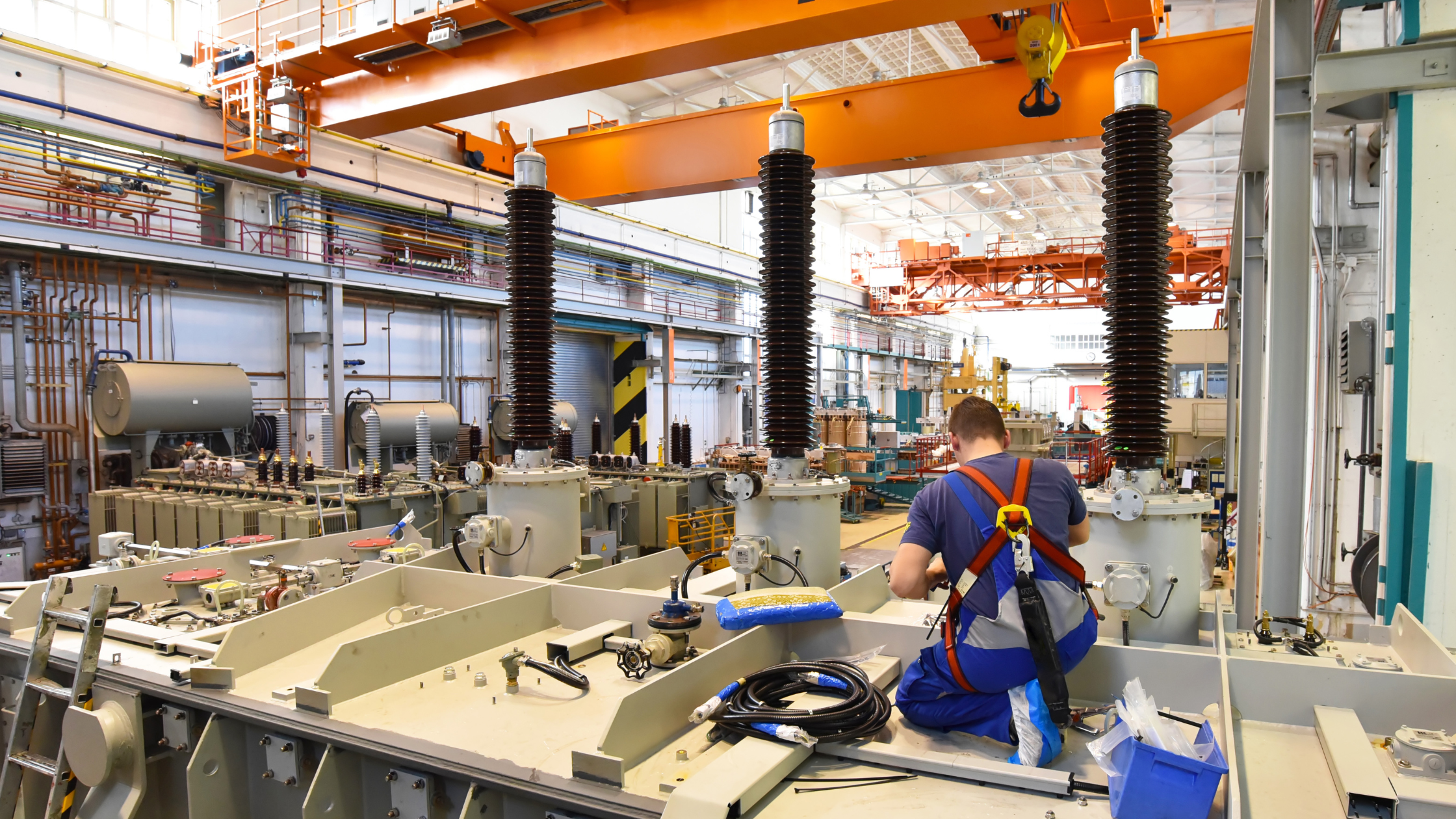 A worker in a harness works to assemble what appears to be equipment for a power substation in a manufacturing facility.